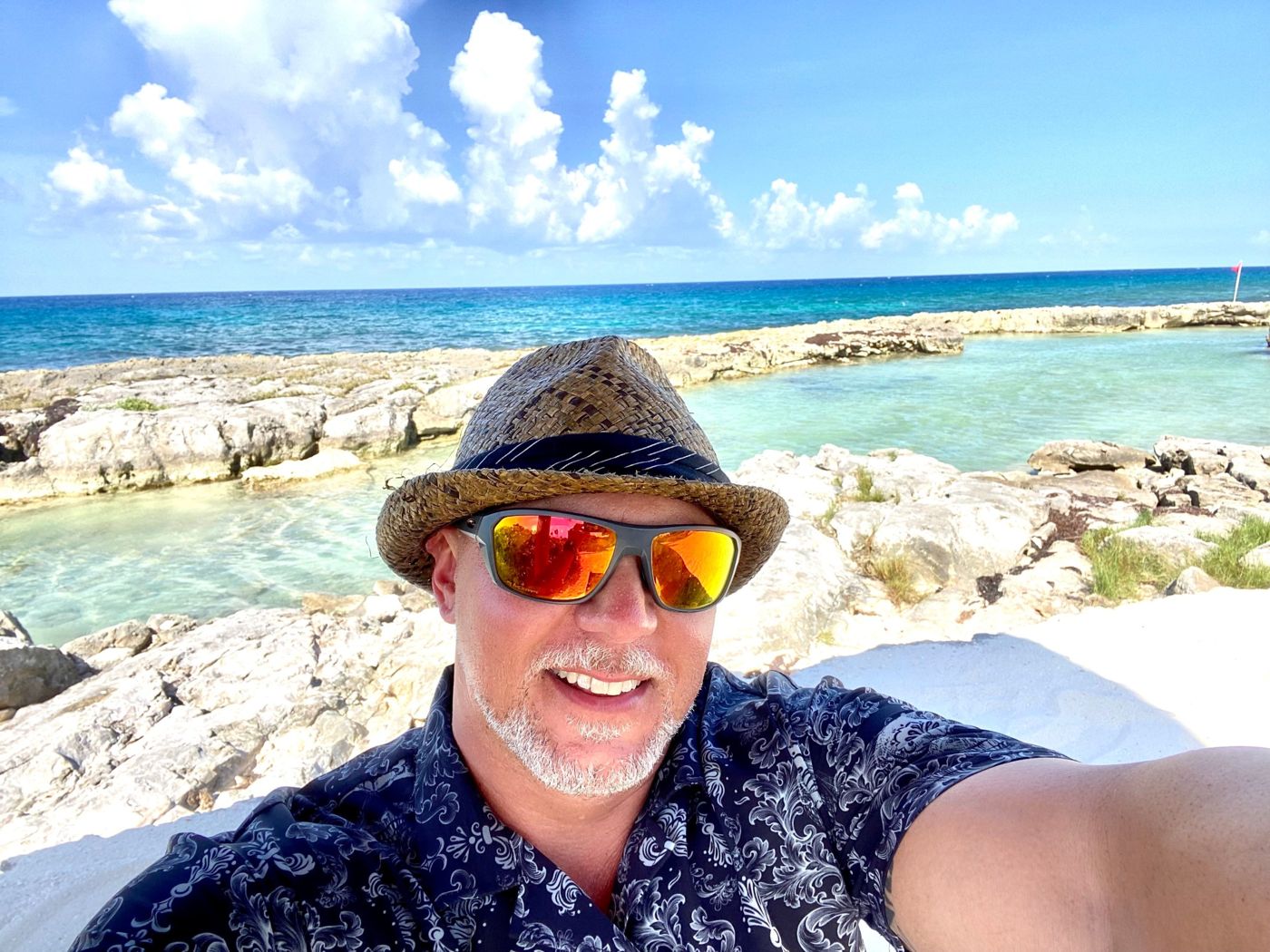 DJ Alex smiling in a straw hat and orange sunglasses at the beach